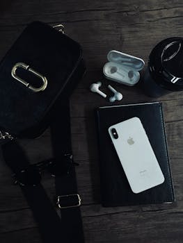 Flat lay of black and white tech accessories including a smartphone, earbuds, and notebook on a wooden surface.