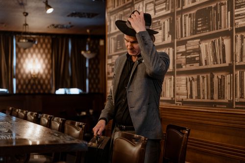 Free A man in a suit and hat adjusts his look while holding a briefcase in a vintage-style room. Stock Photo