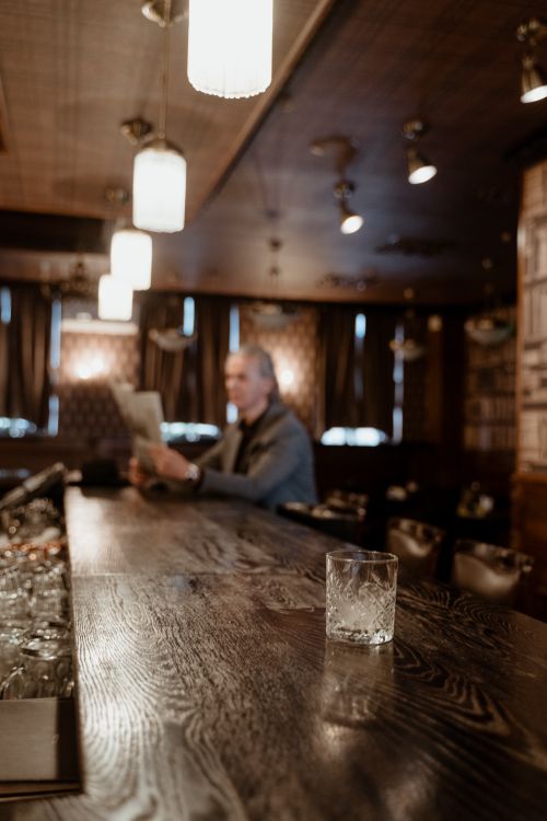 Free Warm bar scene with glass on wooden counter and person reading newspaper. Stock Photo