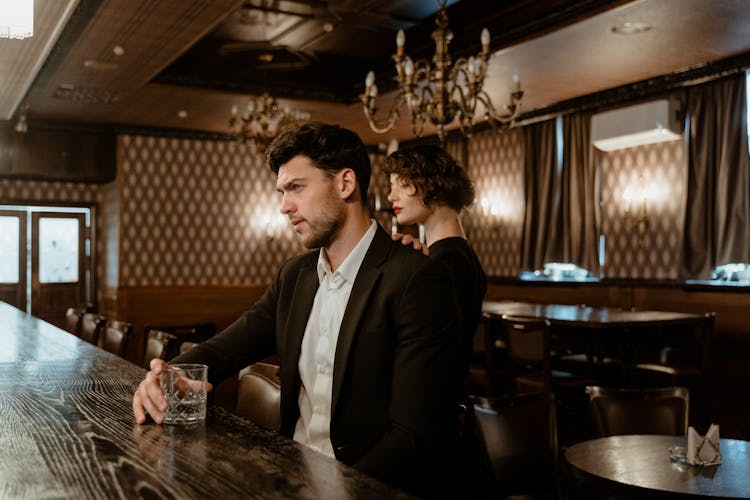 Man In Black Suit Sitting On A Bar Holding A Glass Of Whiskey Beside A Woman In Black Dress