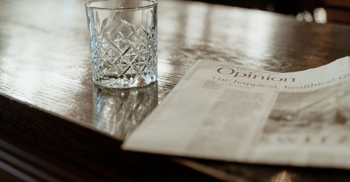 A close-up of a crystal drinking glass and newspaper on a polished wooden bar counter.