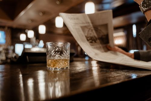Free A close-up of a whiskey glass on a bar counter with a person reading a newspaper in the background. Stock Photo
