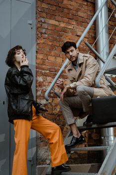 Two fashionable adults smoke and pose on a metal staircase against a brick wall.