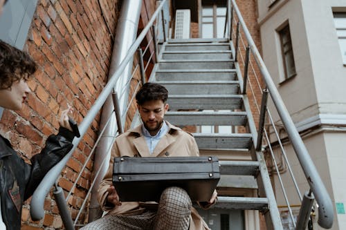 Free Man in Beige Coat Sitting on Metal Stairs Opening a Black Suitcase  Stock Photo