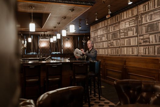 A middle-aged man reads a newspaper in a warm, vintage-style bar filled with books.