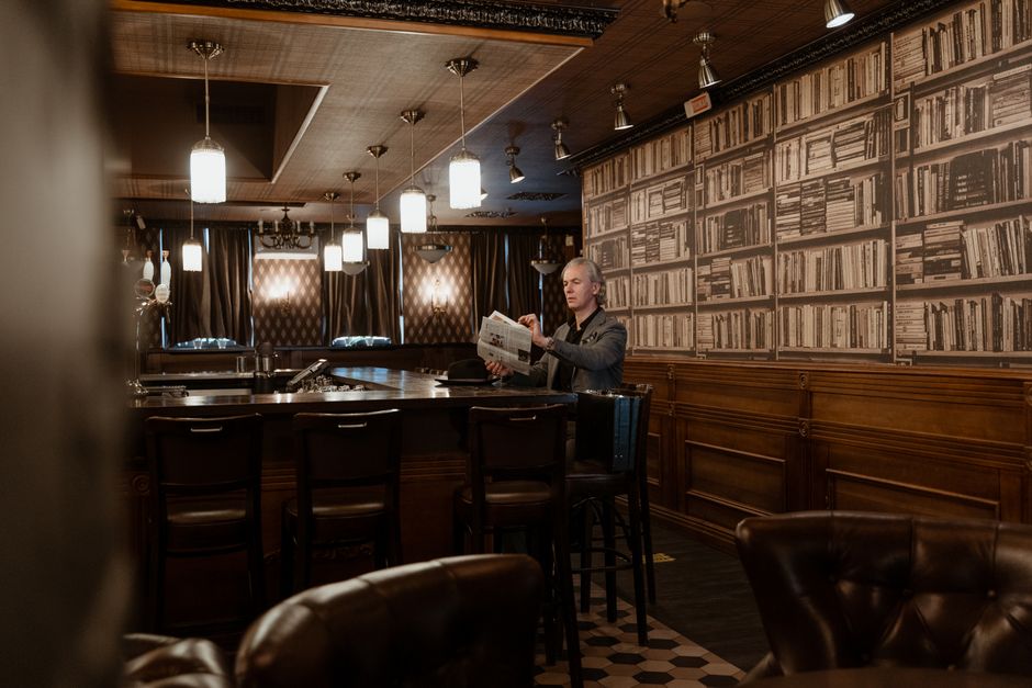 A middle-aged man reads a newspaper in a warm, vintage-style bar filled with books.