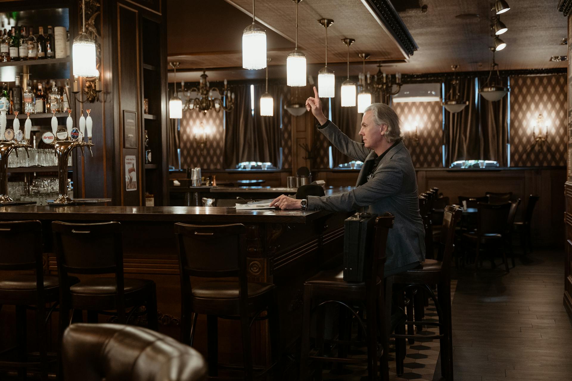 A man in a suit signals for service at a vintage-style bar, indoors atmosphere.