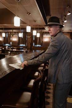 Man in gray suit and hat at a vintage bar, reading a newspaper in a retro setting.
