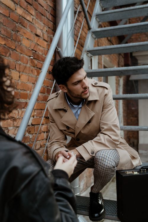 Free Man in a trench coat sitting on outdoor metal stairs, engaged in conversation. Stock Photo