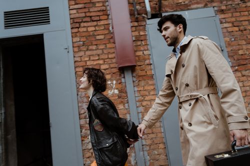 Free A couple walking hand in hand outdoors, the man in a trench coat and the woman in a leather jacket, smiling and stylish. Stock Photo