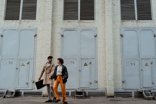 Free A fashionable couple holding hands while walking next to an industrial building with large doors. Stock Photo