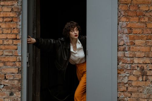 Free Woman in retro style leaning through a doorway in a brick wall, wearing a leather jacket. Stock Photo