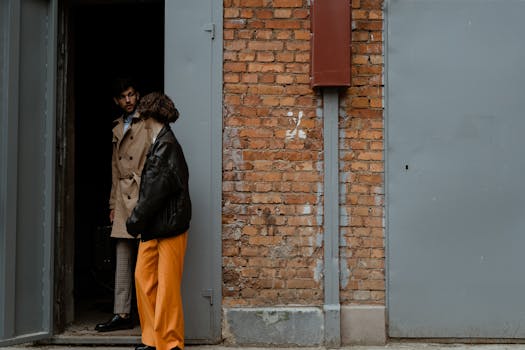 A fashionable couple stands by a brick wall, engaging at a doorway, showcasing urban style.