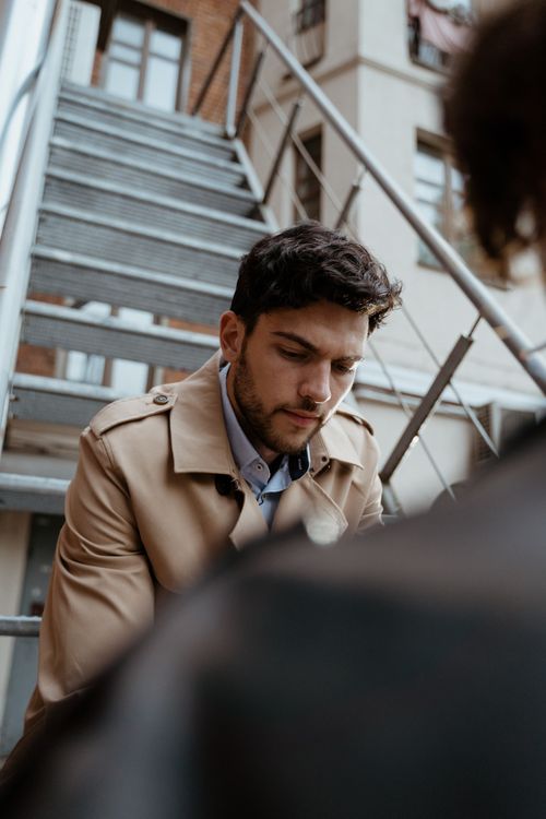 Free Portrait of a fashionable man in a brown coat sitting on metal stairs, showcasing thoughtful expression. Stock Photo