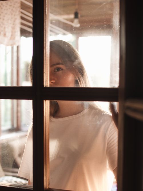 Free A thoughtful woman stands behind a sunlit window, creating a serene atmosphere. Stock Photo