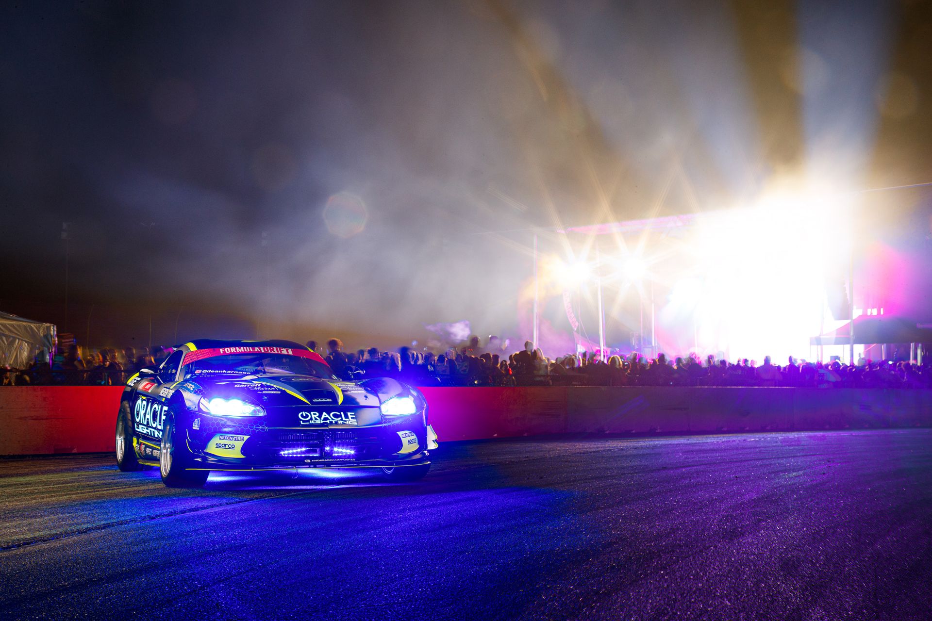 Exciting nighttime motorsport event featuring a race car on a well-lit track with vibrant lights and a cheering crowd.
