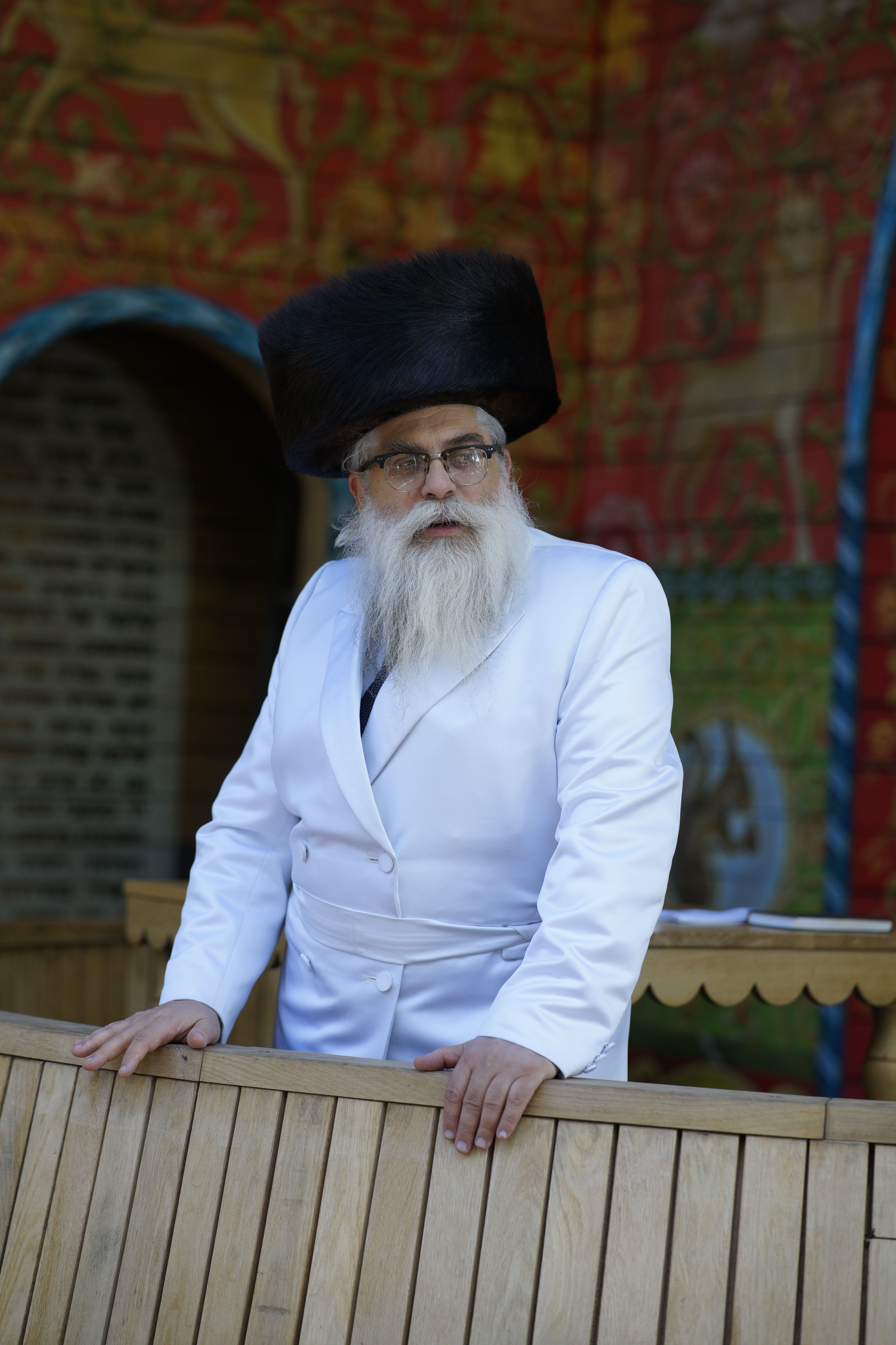 Elderly bearded man in traditional attire leaning on wooden railing, captured outdoors.