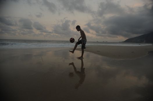 A person kicks a soccer ball on the beach with the ocean and clouds in the background at dusk.