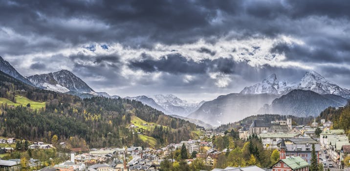 A picturesque alpine town nestled in a valley with majestic snowy mountains under a cloudy sky.