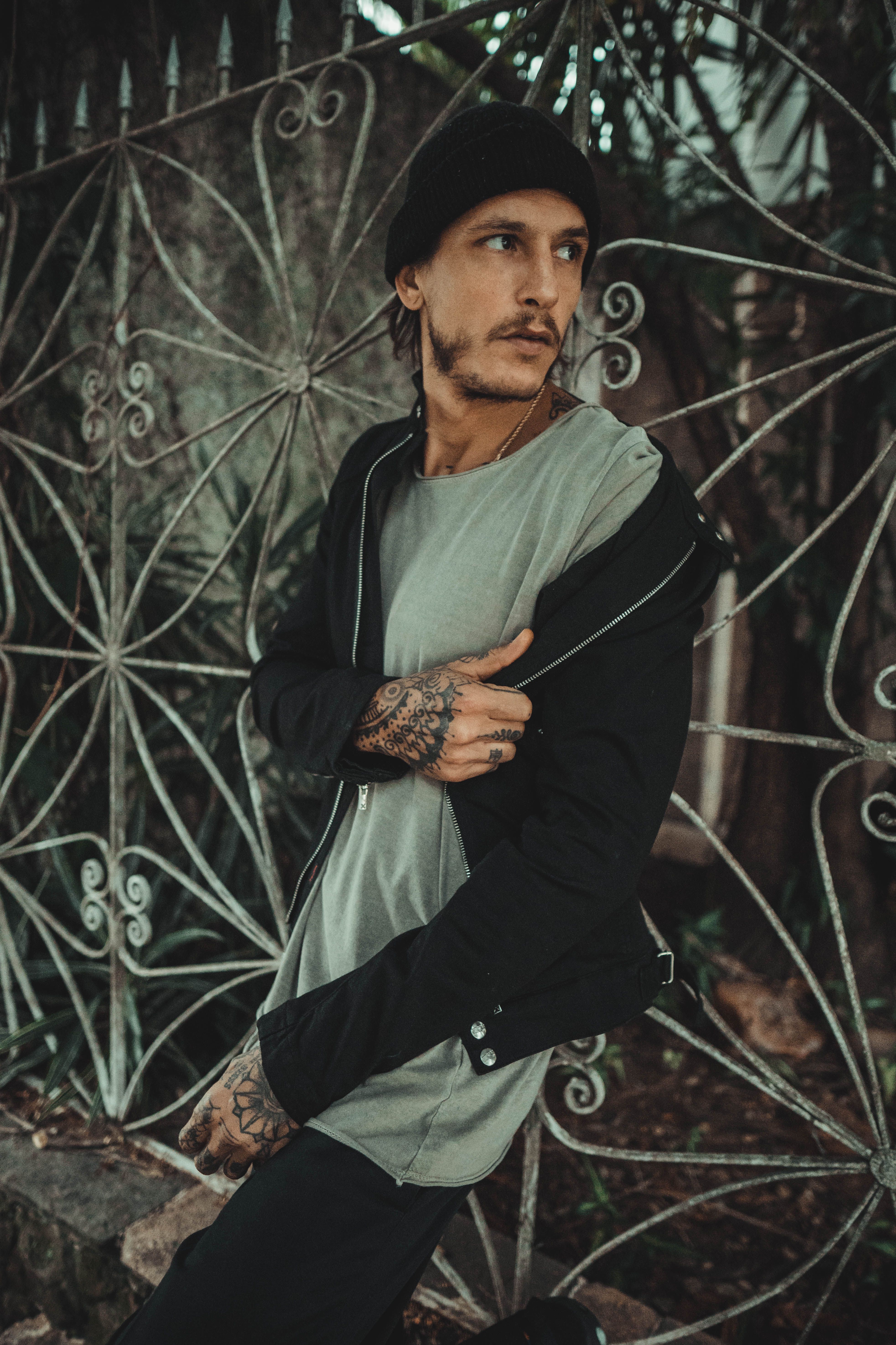 A stylish tattooed man in a black jacket poses against a decorative gate in São Paulo.