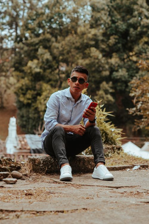 Free Stylish young man wearing sunglasses and using smartphone while sitting outdoors in a park setting. Stock Photo