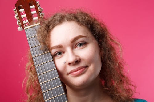 Close-Up Shot of a Curly-Haired Woman Holding an Acoustic Guitar on Pink Background