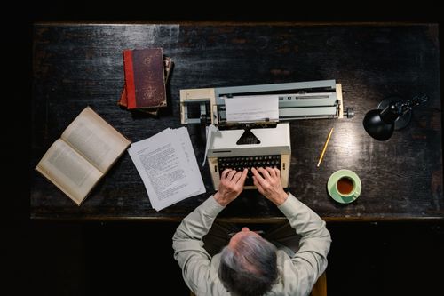 Free Senior man using typewriter at wooden desk surrounded by books and tea. Stock Photo