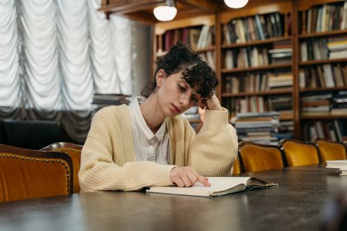 Free A young woman reading a book while studying indoors in a university library setting. Stock Photo