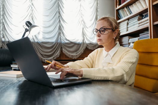 Mature woman in glasses working intently on a laptop at a library desk.