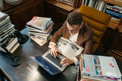 Free A young man studies in a cozy library setting, using a laptop and books at a wooden desk. Stock Photo