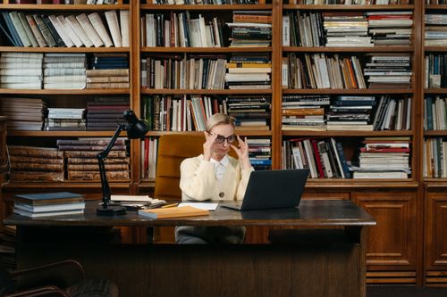 Free A woman working on her laptop in a library room with shelves full of books. Stock Photo