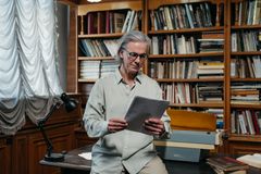 A senior man with glasses reads papers in a library filled with books, creating a scholarly atmosphere.