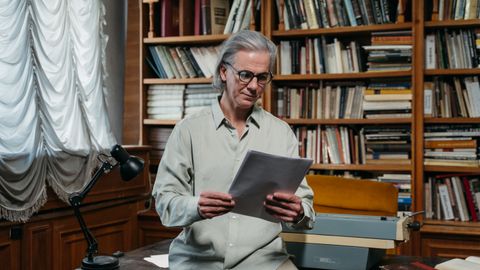 A senior man with glasses reads papers in a library filled with books, creating a scholarly atmosphere.