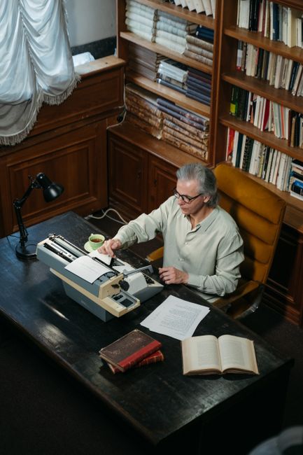 Gray-haired man typing on a typewriter in a cozy library setting with bookshelves and warm lighting. Gray-haired man typing on a typewriter in a cozy library setting with bookshelves and warm lighting.