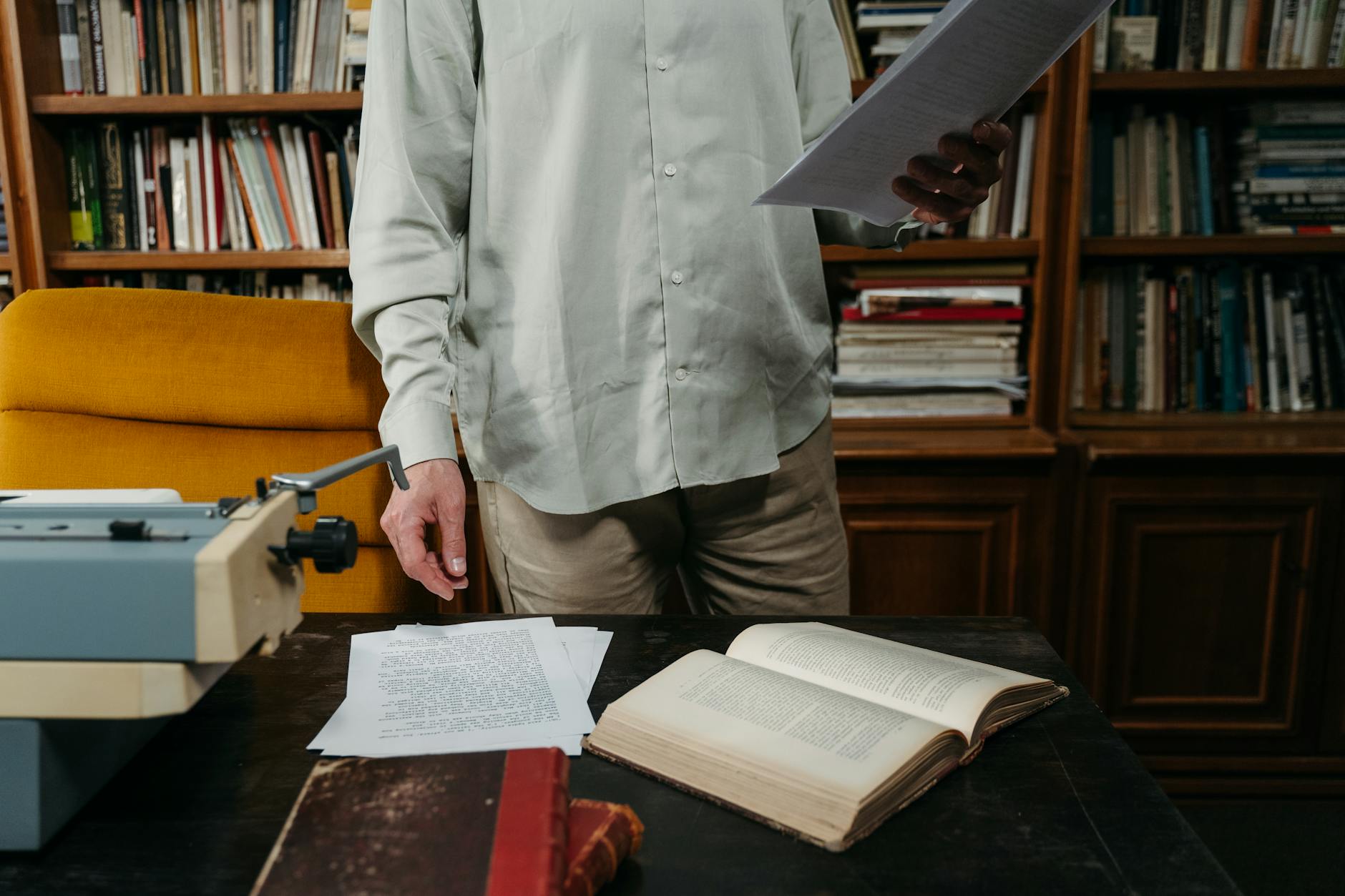Man reviewing documents in a cozy library with books, papers, and typewriter.