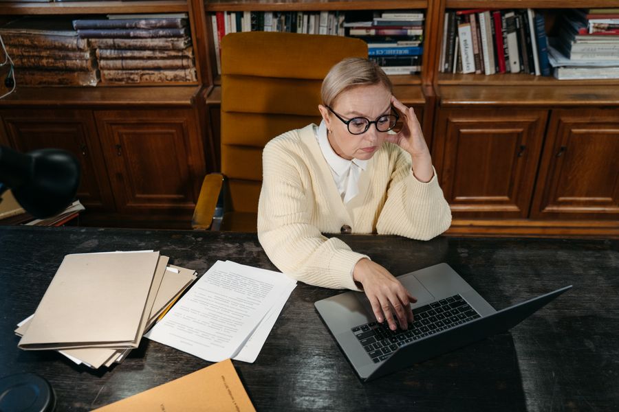 Mature woman wearing eyeglasses working on a laptop at a desk in a library, highlighting learning and concentration.