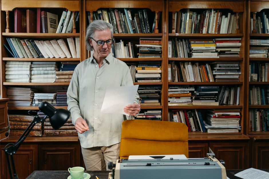 Elderly man reading a document in a cozy home library with bookshelves.