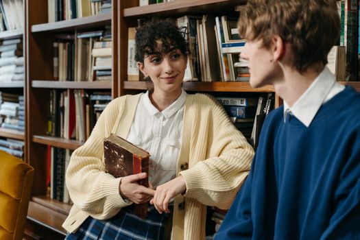 Two students have a cheerful conversation in a library setting with bookshelves in the background.