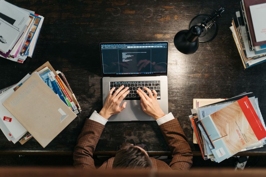 A person typing on a laptop at a cluttered desk with books and documents around.
