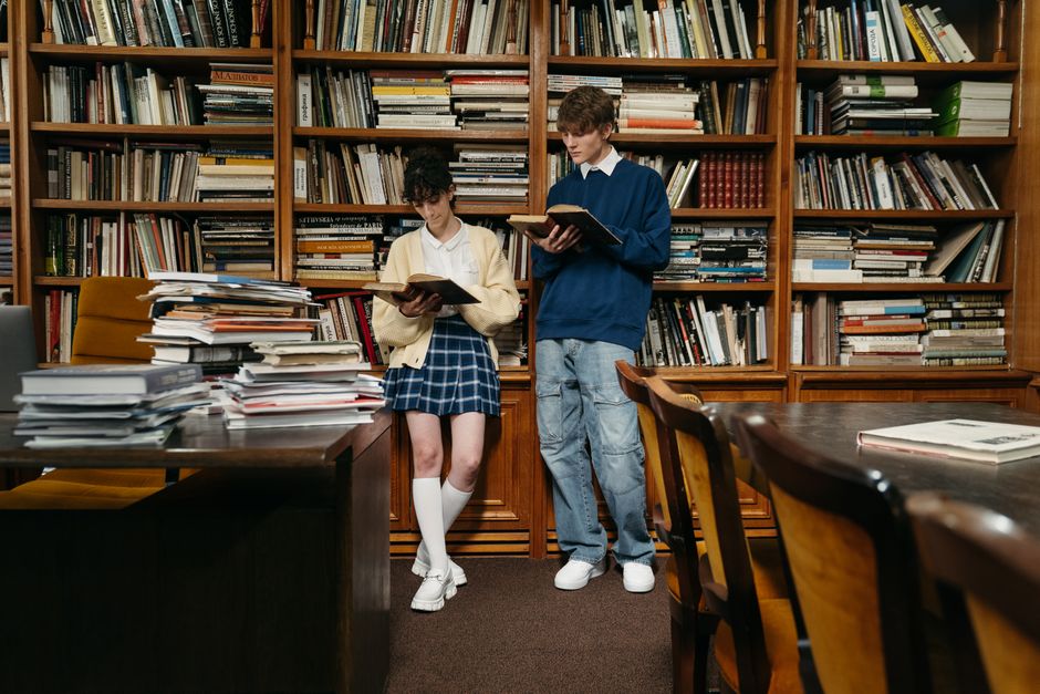 Two students reading books in a cozy library setting with wooden bookshelves.