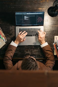 A person in a suit types on a laptop keyboard at a wooden desk, viewed from above.