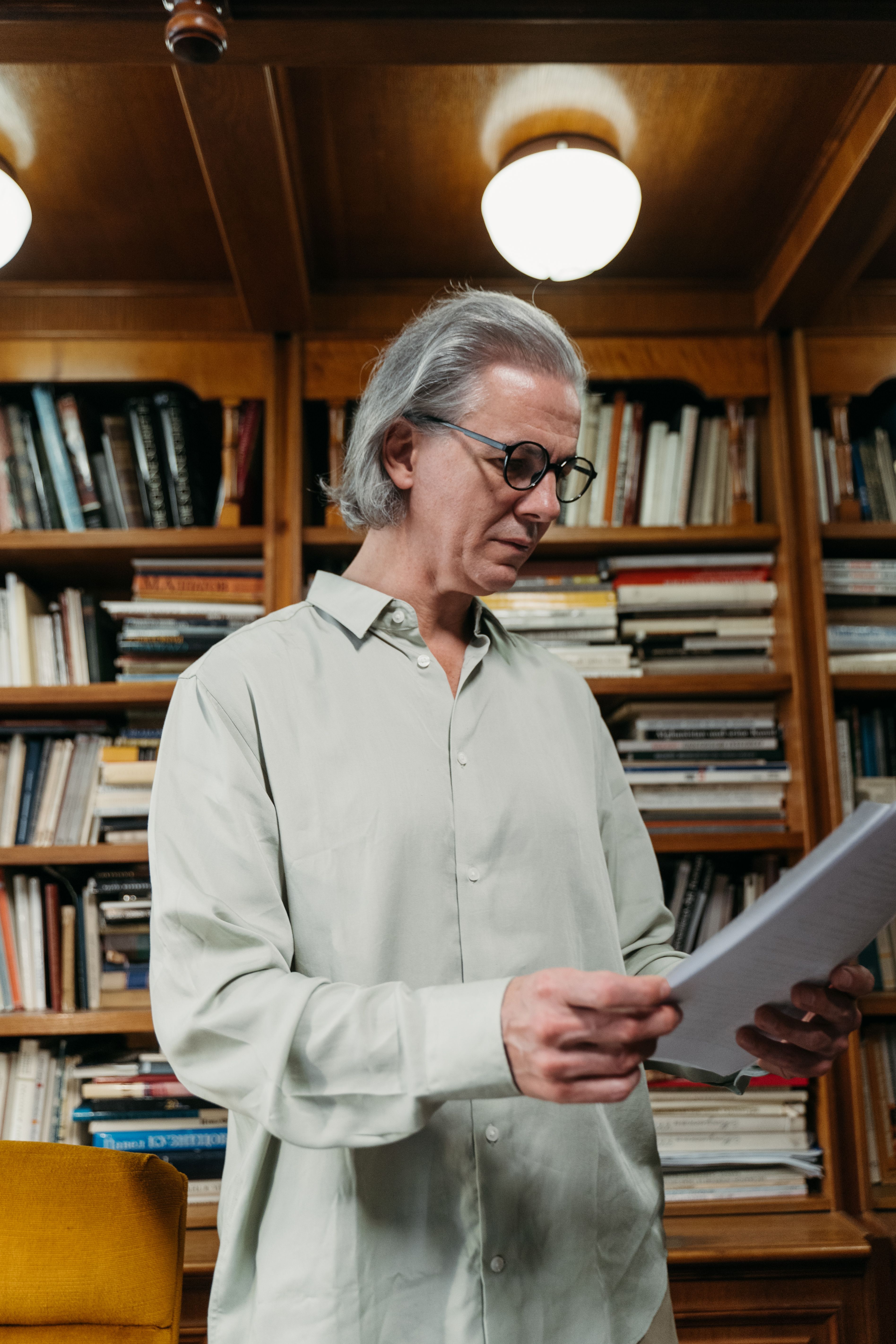 Senior man with eyeglasses reading papers in a library surrounded by bookshelves.