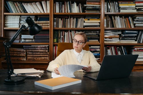 Free A senior woman focuses on reading documents at a desk in a library, surrounded by books and a laptop. Stock Photo