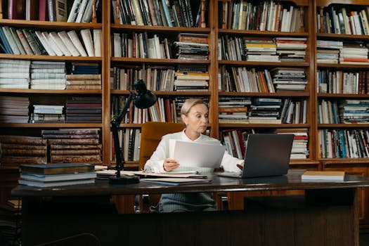 Focused woman working on a laptop in a library surrounded by books, depicting remote work and study.