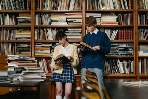 A Man and a Woman Reading Books while Leaning on a Bookcase