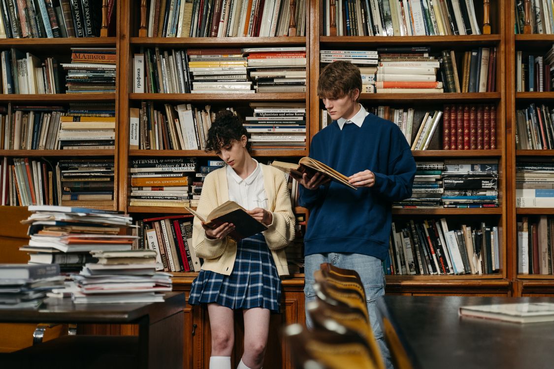 Free A Man and a Woman Reading Books while Leaning on a Bookcase Stock Photo