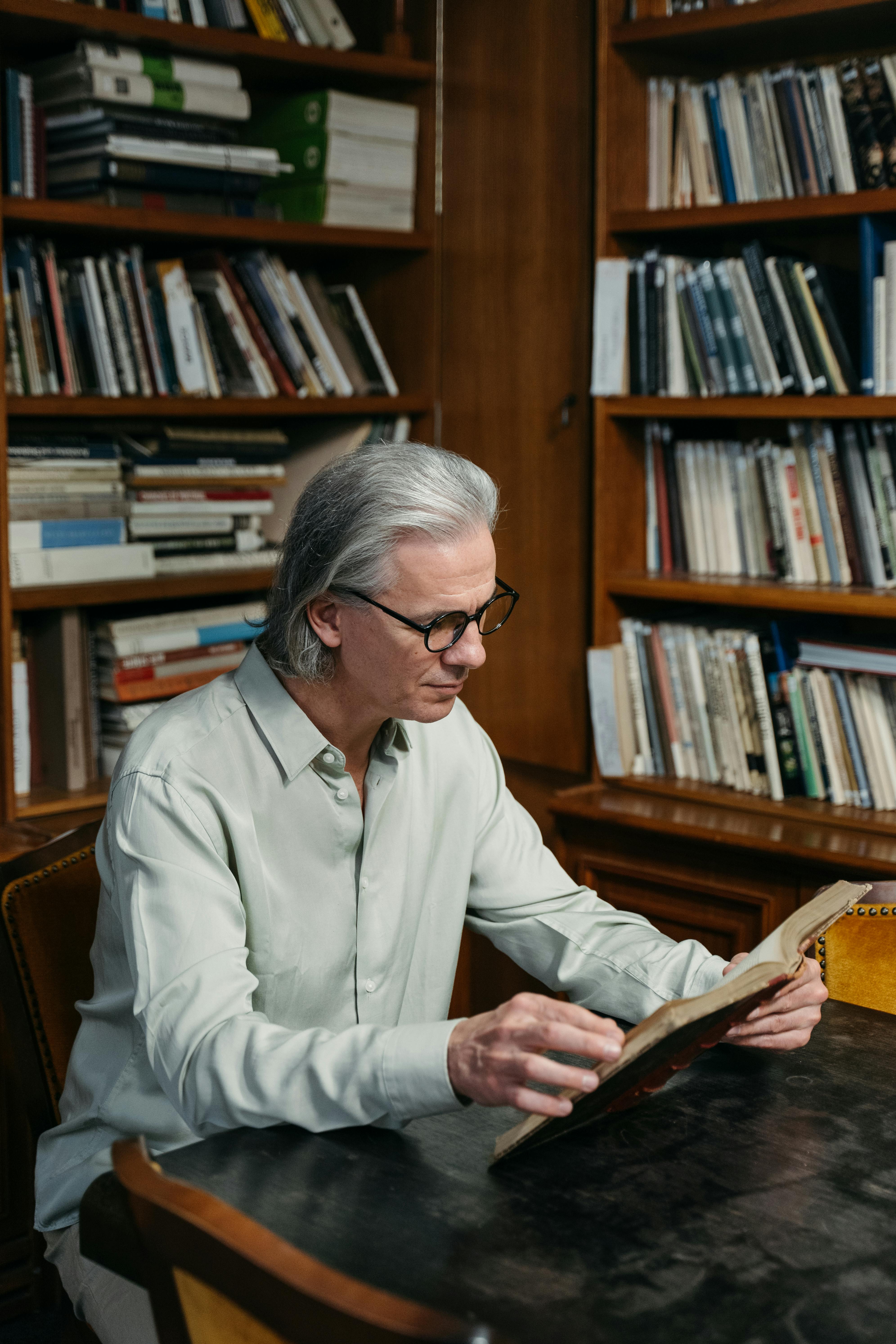 Senior man in a light shirt reading in a cozy library with numerous bookshelves.