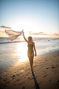 Caucasian woman in swimsuit and shawl walking on a serene beach during sunset, capturing a moment of relaxation.
