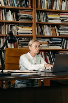 An elderly woman wearing eyeglasses types on a laptop in a library, surrounded by books.