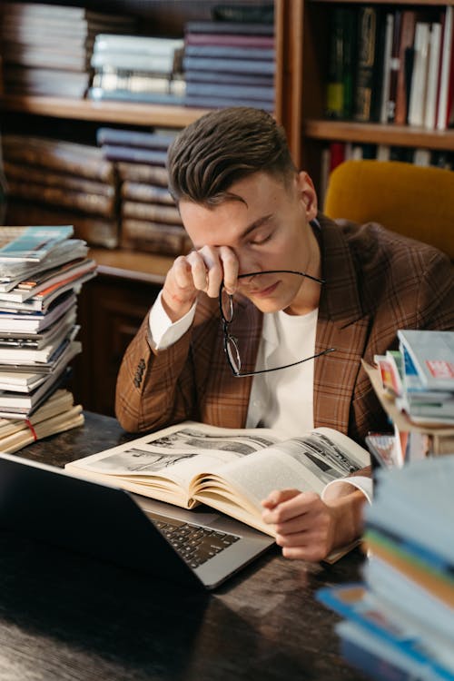 Free Tired Student Rubbing His Eyes while Sitting in front of a Laptop Surrounded by Piles of Books  Stock Photo